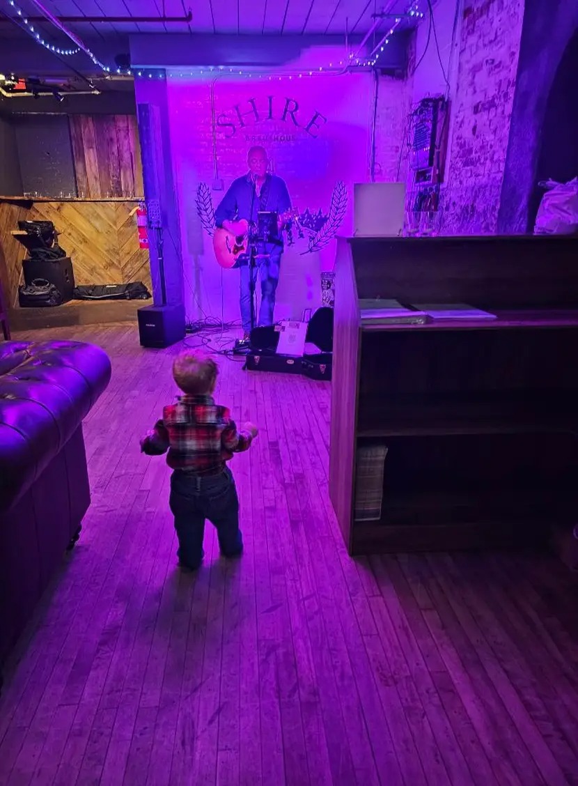 A child holding a teddy bear walks toward an adult in a purple-lit room.