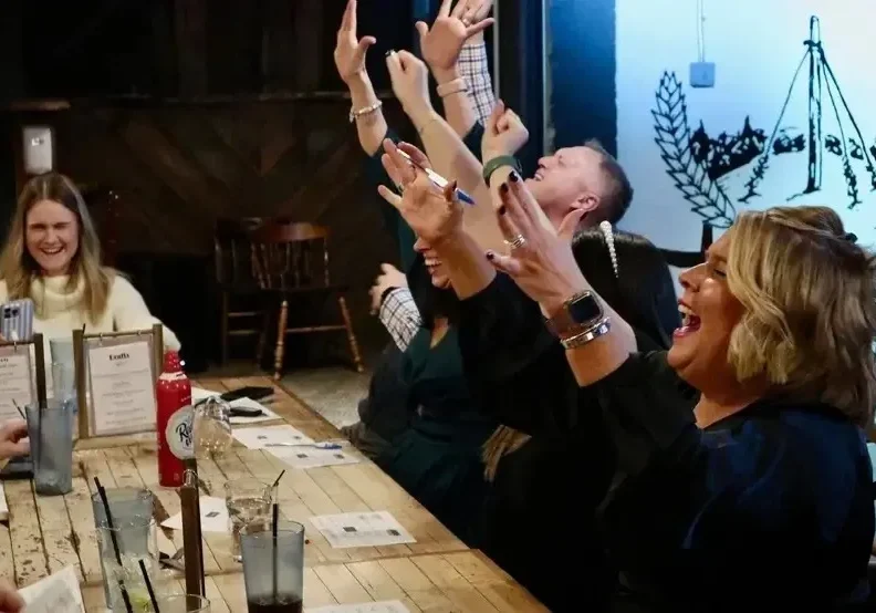People raising hands joyfully around a wooden table in a lively setting.